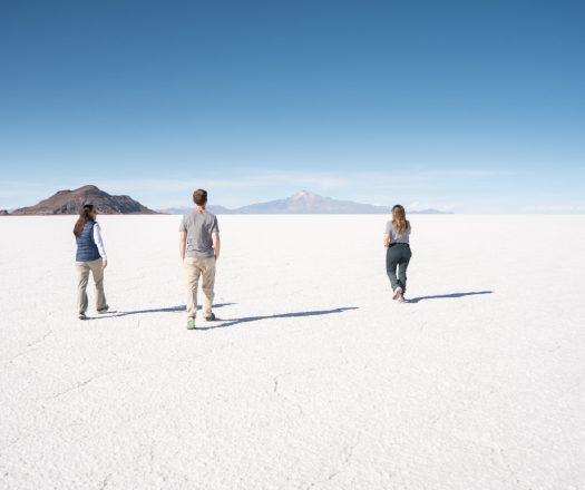 Three people walking across a vast white salt flat toward distant mountains.