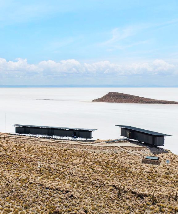 Minimalist Explora lodge structures overlooking vast white salt flats, with volcanic terrain under a clear desert sky in Bolivia.