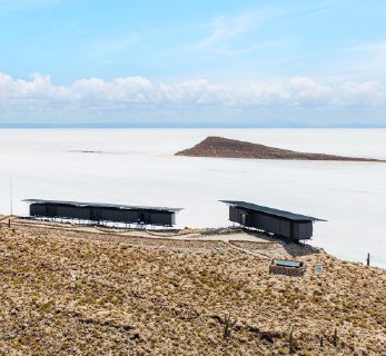 Minimalist Explora lodge structures overlooking vast white salt flats, with volcanic terrain under a clear desert sky in Bolivia.