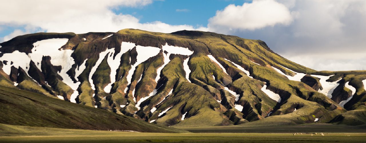 http://A%20wide%20view%20of%20rolling%20green%20mountains%20marked%20with%20streaks%20of%20melting%20snow%20under%20a%20dramatic,%20cloud-filled%20sky.