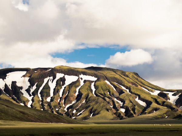 http://A%20wide%20view%20of%20rolling%20green%20mountains%20marked%20with%20streaks%20of%20melting%20snow%20under%20a%20dramatic,%20cloud-filled%20sky.
