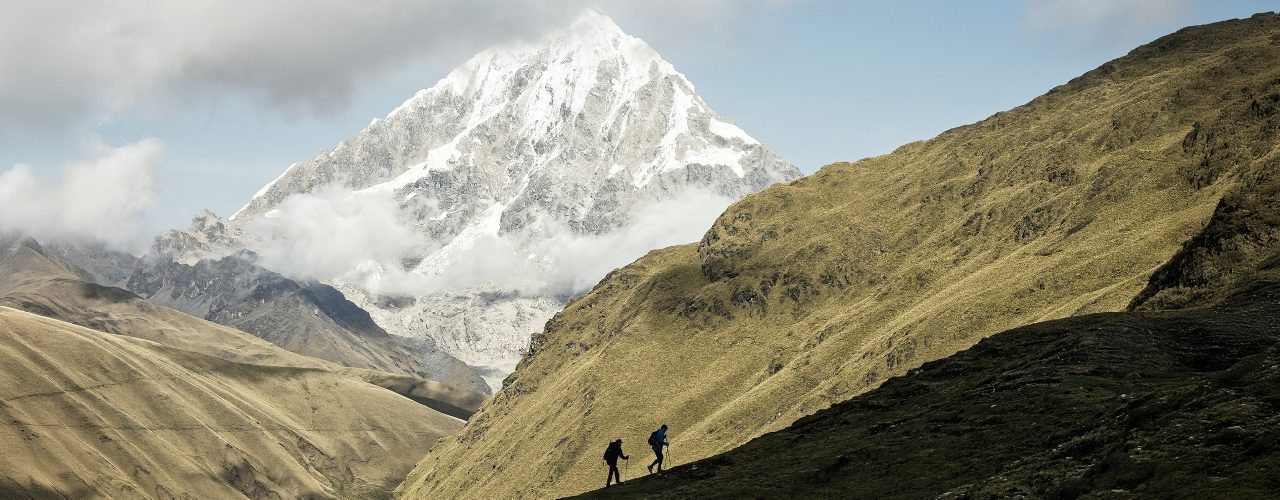 http://Two%20hikers%20walk%20along%20a%20grassy%20hillside,%20dwarfed%20by%20a%20towering,%20snow-covered%20mountain%20partially%20wrapped%20in%20clouds.