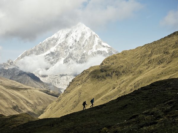 http://Two%20hikers%20walk%20along%20a%20grassy%20hillside,%20dwarfed%20by%20a%20towering,%20snow-covered%20mountain%20partially%20wrapped%20in%20clouds.