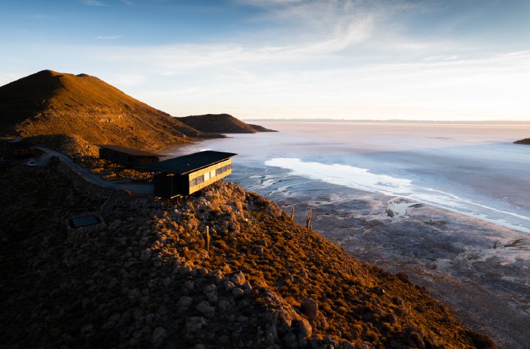 A modern lodge perched on a rocky hillside overlooks a vast salt flat glowing in early morning light.