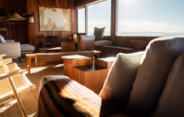 A cozy wooden lounge with sofas and large windows framing an open desert and distant mountains.