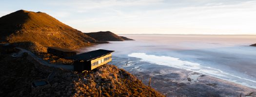 A modern lodge clings to a hillside overlooking vast salt flats in golden sunset light.