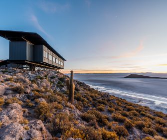 A modern building sits on a rocky hillside overlooking a vast lake or salt flat, surrounded by sparse desert vegetation and lit by warm sunset light.