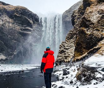 Expedition traveler standing before Skógafoss waterfall in winter, Iceland — powerful cascading falls and snow-covered basalt cliffs