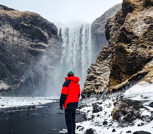 Expedition traveler standing before Skógafoss waterfall in winter, Iceland — powerful cascading falls and snow-covered basalt cliffs